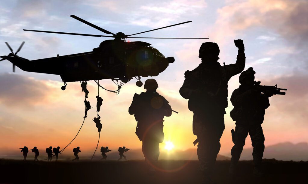 A backlit image of three soldiers walking towards the camera, with a helicopter releasing more behind.